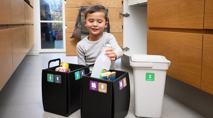 Dreng sidder på køkkengulv og sorterer plastdunk i affaldsspand til genanvendelse